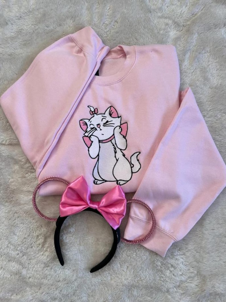 K&ouml;nnte beinhalten: Hellrosa Sweatshirt mit gestickter wei&szlig;er Katze und rosa Akzenten. Ein passendes rosa Schleifen-Stirnband mit schwarzem Band und glitzernden Ohren liegt neben dem Sweatshirt. Die Katze ist aus Aristocats.