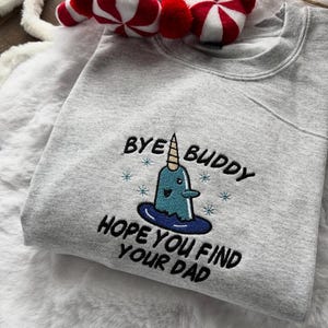 Pode incluir: Um moletom cinza mesclado com a frase bordada "Bye Buddy, Hope You Find Your Dad" e um desenho de narval. O narval azul tem um chifre branco e flocos de neve azuis. Uma decoração listrada em vermelho e branco é visível no fundo.