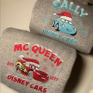 May include: Two gray sweatshirts with embroidered designs. One features a blue car with a Santa hat, the name "SALLY", and "DISNEY CARS". The other displays a red car with a Santa hat, the name "MC QUEEN", and "DISNEY CARS".