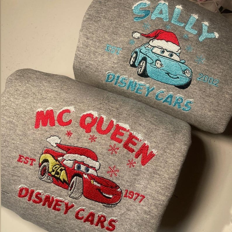 May include: Two gray sweatshirts with embroidered designs. One features a blue car with a Santa hat, the name "SALLY", and "DISNEY CARS". The other displays a red car with a Santa hat, the name "MC QUEEN", and "DISNEY CARS".