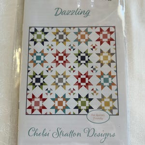 以下が含まれることがあります： Chelsi Stratton Designsの「Dazzling」というタイトルのキルトパターンのパッケージ。パターンは、赤、緑、青、黄色など、さまざまな色の星のキルトデザインを特徴としています。パッケージには「Fat Quarter Friendly」と表示されています。