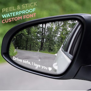 May include: A car side mirror with the text "Drive safe, I love you" and a heart graphic. The mirror reflects a road winding through a forest. Above the mirror is the text "PEEL & STICK WATERPROOF CUSTOM FONT".