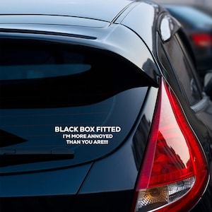 May include: A black car with a white decal on the rear window. The decal reads "BLACK BOX FITTED I'M MORE ANNOYED THAN YOU ARE!!!" The car's taillight is red and white.