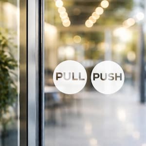 May include: Glass door with two white circular signs. One sign says "PULL" and the other says "PUSH" in gray text. The door is framed by a silver metal border. The background is blurred, showing a bright interior.