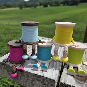 May include: Five small, articulated figurines shaped like spools of thread in bright colors including light blue, yellow, pink, teal, and lime green. Each spool has jointed arms and legs with rounded feet, allowing them to sit upright.