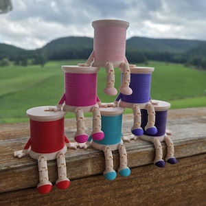 May include: A pyramid stack of six colorful thread spools, each featuring articulated light-colored arms and legs with matching colored feet. The spools are arranged in three tiers, displaying vibrant shades of red, pink, purple, and blue thread.