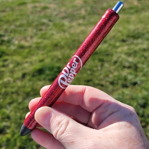 May include: A red glitter pen with a black tip and a silver top. The pen has the words "Dr Pepper" printed on it in white.