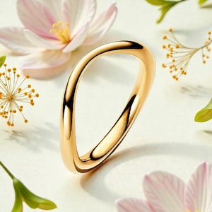 May include: A gold ring with a distinctive, flowing design. The ring is set against a backdrop of white and pink flowers and green foliage. The ring's smooth, curved shape is the focal point, creating an elegant and modern aesthetic.