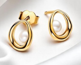 May include: A pair of gold-colored stud earrings. Each earring features a small, white pearl set within an oval-shaped, open-frame design. The earrings are displayed on a white, silky fabric, highlighting their polished finish and elegant simplicity.