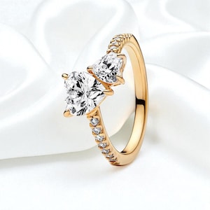 May include: A gold-colored ring featuring two heart-shaped, clear gemstones. The ring band is adorned with small, clear gemstones. The ring is set against a white, textured background.