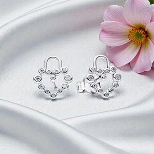 May include: Silver stud earrings with a heart-shaped design. Each earring has small, clear gemstones set around the heart, with a small loop at the top. The earrings are displayed on a white, silky surface. A pink flower is in the background.