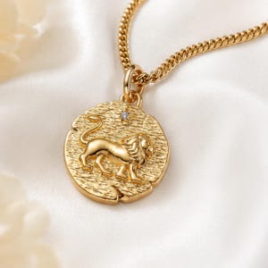 May include: A gold-toned necklace featuring a round pendant with a textured surface and a raised lion figure in the center. A small, clear, star-shaped stone is set above the lion. The pendant hangs from a matching gold-toned curb chain link.