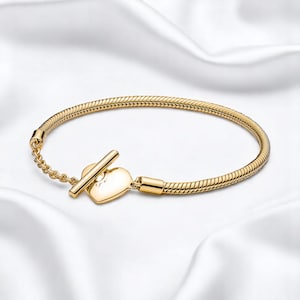 May include: A gold bracelet with a heart-shaped clasp. The bracelet has a snake chain design and a heart-shaped clasp with a small, sparkling detail. The clasp is secured with a bar closure and a small chain.