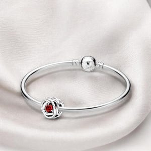 May include: A silver bracelet with a round clasp and a charm. The charm is a silver knot design with a small, round, red gemstone in the center. The bracelet is displayed on a white, silky surface.