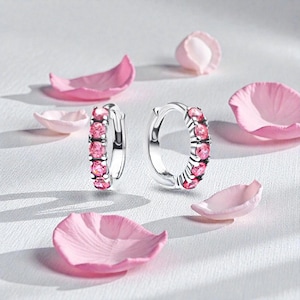 May include: A pair of silver hoop earrings with a row of pink gemstones. The earrings are displayed on a white surface, surrounded by pink flower petals. The gemstones are a vibrant pink color, adding a touch of elegance.