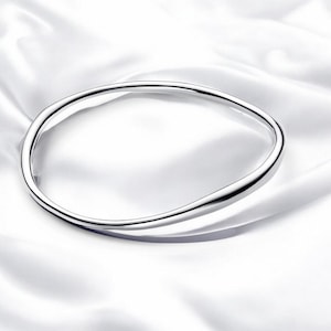 Puede incluir: Una pulsera de plata con un diseño liso y curvado. La pulsera tiene forma ovalada y parece estar hecha de metal pulido. El fondo es una tela blanca y sedosa, que resalta la superficie reflectante de la pulsera.