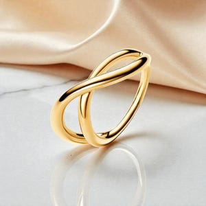 Op de afbeelding: Een goudkleurige ring met een oneindigheidssymboolontwerp. De ring is gemaakt van een glanzend, reflecterend metaal en staat tegen een zachte, neutrale achtergrond. Het ontwerp van de ring heeft twee in elkaar grijpende lussen.