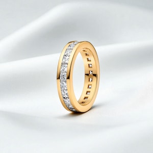May include: A gold ring with a continuous row of square-cut, clear gemstones. The ring has a polished, yellow-gold band. The gemstones are set in a channel setting, creating a seamless, sparkling appearance. The ring is displayed on a white background.