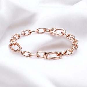 May include: Rose gold chain link bracelet. The bracelet features a series of interconnected oval and rectangular links. The clasp is a matching rose gold colour. The bracelet is displayed on a white, silky background.