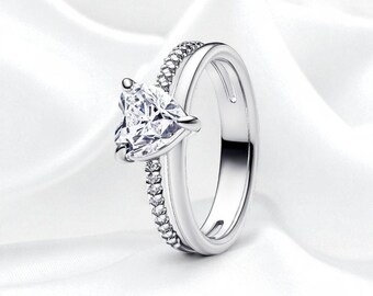 May include: A silver ring with a pear-shaped, clear gemstone as the focal point. The ring has a split band design, with one side adorned with small, clear stones. The ring is set against a white background.