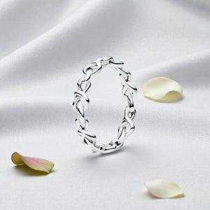 May include: A silver-colored ring with a woven design, resting on a white satin surface. The ring's intricate pattern is highlighted by the light, and the image includes scattered flower petals.