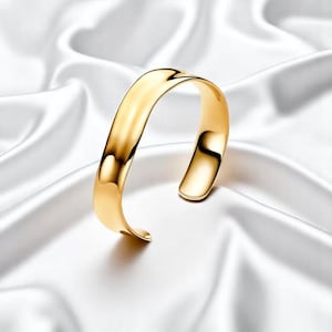 Puede incluir: Un brazalete dorado con un diseño curvo. El brazalete abierto está hecho de un metal brillante y reflectante. La joya se exhibe sobre una tela blanca y sedosa.
