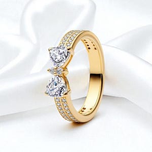 Op de afbeelding: Een goudkleurige ring met een vlinderdasontwerp. De ring heeft twee hartvormige heldere stenen en is bezet met kleine, heldere stenen langs de band. De ring staat tegen een witte achtergrond.
