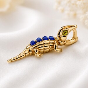 Puede incluir: Un colgante de cocodrilo de metal dorado con un cuerpo texturizado y segmentado, cuatro piedras piramidales de color azul intenso en el lomo y una piedra de color verde brillante como ojo. La pieza incluye una anilla articulada en la cabeza.