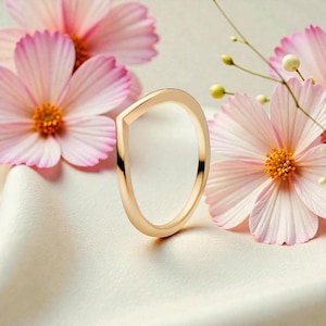 May include: A gold-colored ring with a unique, angular design. The ring is set against a backdrop of soft pink cosmos flowers and a white, silky fabric. The ring's shape is modern and minimalist, with a flat top and curved sides.