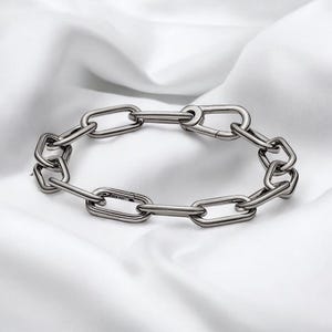 Puede incluir: Un brazalete de eslabones plateado. El brazalete presenta una serie de eslabones ovalados alargados conectados entre sí. Los eslabones son lisos y tienen un acabado pulido. El brazalete se muestra sobre una superficie blanca y texturizada.