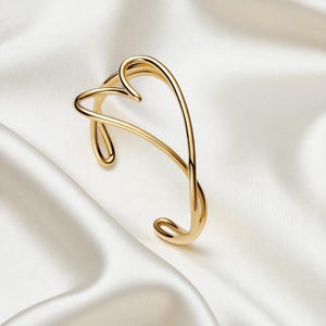Könnte beinhalten: Ein goldfarbenes Armreif-Armband mit herzförmigem Design. Das Armband besteht aus einem durchgehenden, gebogenen Metallband, das die Form eines Herzens bildet. Es wird auf einer weißen, seidigen Oberfläche präsentiert.