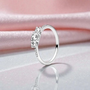 May include: A silver ring with three clear, round gemstones set in a row. The band is thin and also features small, clear stones. The ring is displayed against a soft pink background, reflecting light.