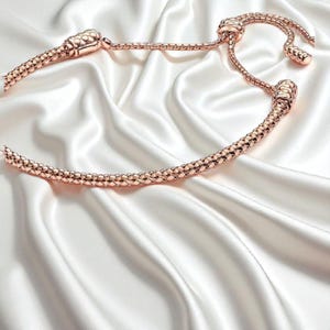 Puede incluir: Un collar de oro rosa con un diseño único, sobre una superficie de satén blanco. El collar presenta una cadena texturizada y cierres decorativos. El diseño general es elegante y sofisticado.