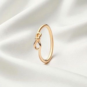 Op de afbeelding: Een goudkleurige ring met een strikvormig ontwerp aan de voorkant. De ring is geplaatst tegen een witte, zijdeachtige achtergrond. De strik is een delicaat detail en voegt een vleugje elegantie toe aan het sieraad.