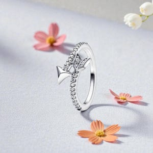 Könnte beinhalten: Ein silberner Ring mit floralem Design. Der Ring hat eine Reihe kleiner, klarer Steine entlang des Bandes und ein dekoratives Blumenelement mit klaren Steinen. Der Ring ist vor einem weichen, hellen Hintergrund mit verstreuten rosa und orangefarbenen Blumen platziert.