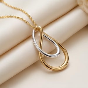May include: A necklace featuring a pendant composed of two interlocking teardrop-shaped loops, one in polished gold and one in silver, suspended from a delicate gold chain. The design creates a continuous, fluid shape with a smooth finish.