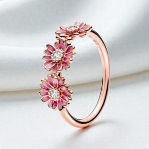 May include: Rose gold ring featuring three pink daisy flower designs with white gemstone centers. The flowers are enameled with a darker pink outline. The ring is set against a white background.