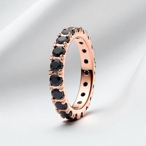 May include: A rose gold ring set with a continuous line of round, black gemstones. The band has a textured design, and the gemstones are closely set, forming a full circle of dark sparkle. The ring is shown against a soft, white background.