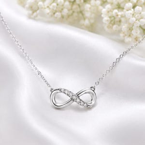 Pode incluir: Um colar prateado com um pingente em forma de símbolo do infinito. O pingente é adornado com pequenas pedras preciosas transparentes. O colar repousa sobre um tecido branco e sedoso, com flores brancas ao fundo.