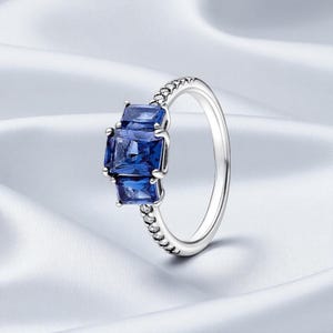 Op de afbeelding: Een zilveren ring met drie vierkante, diepblauwe edelstenen. De edelstenen zijn in een rij geplaatst, met een kralenontwerp langs de band. De ring wordt getoond op een witte, zijdeachtige achtergrond.