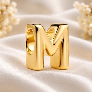 Op de afbeelding: Een gepolijste goudkleurige metalen kraal in de vorm van de hoofdletter M. De kraal heeft een glad, afgerond oppervlak met een reflecterende afwerking en een centrale verticale opening om op een koord, ketting of armband te rijgen.