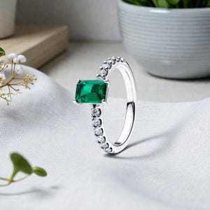 May include: A silver ring with a rectangular, emerald-cut green gemstone as the focal point. The band is adorned with small, round, clear gemstones. The ring is set against a white, textured background, showcasing its elegant design.