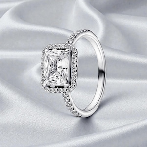 Peut inclure: Une bague en argent ornée d'une grande pierre précieuse rectangulaire et transparente, entourée de petites pierres transparentes. Le corps de la bague est également orné de petites pierres transparentes, créant un effet scintillant. La bague est posée sur un fond blanc doux.