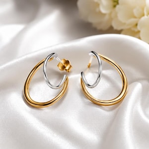 May include: A pair of two-tone hoop earrings featuring a large polished gold-tone oval hoop intertwined with a smaller silver-tone hoop. The earrings include a gold-tone post and a matching butterfly-style earring back for secure fastening.