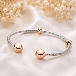 May include: A silver-tone bangle bracelet with rose gold accents. The bracelet features a woven cable design and is adorned with three rose gold spheres and two smaller rose gold beads. The bracelet is displayed on a white satin surface.