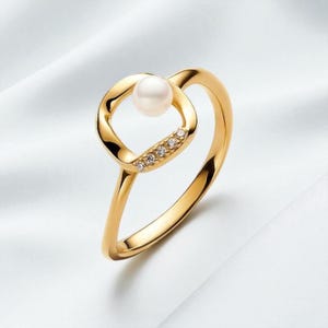 Puede incluir: Un anillo de oro con una perla blanca engastada en un diseño cuadrado. El anillo tiene una banda pulida y está adornado con pequeñas piedras brillantes. El anillo se presenta sobre una superficie blanca texturizada.