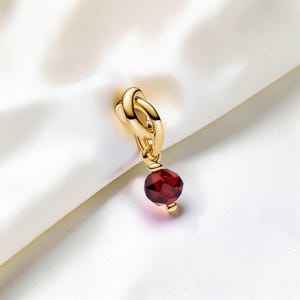 Puede incluir: Un dije de metal dorado con un diseño de nudo entrelazado suspendido de una pequeña anilla. Una cuenta de piedra preciosa de color rojo intenso y facetada cuelga de la parte inferior del nudo, asegurada por una pequeña tapa de metal dorado.