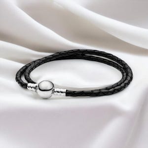 May include: A black braided leather bracelet with a silver clasp. The bracelet is double-stranded and lies on a white, silky surface. The clasp is a round, silver bead with silver accents.
