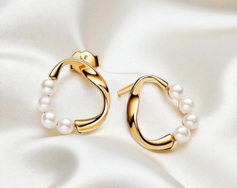 May include: A pair of gold-colored hoop earrings with a curved design. Each earring features a series of small, white pearls. The earrings are displayed on a white, silky fabric, creating a luxurious aesthetic.
