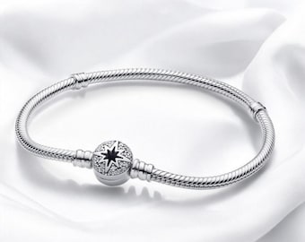 May include: A silver bracelet with a snake chain design. The bracelet features a round charm with a black star design and is encrusted with small, clear crystals. The bracelet is displayed on a white, silky background.